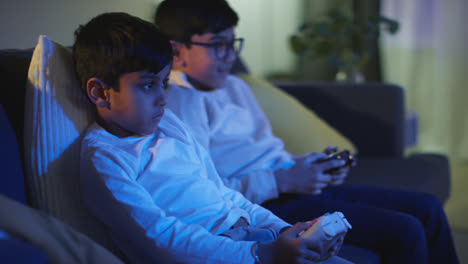 Side-View-Of-Two-Young-Boys-At-Home-Having-Fun-Playing-With-Computer-Games-Console-On-TV-Holding-Controllers-Late-At-Night-1